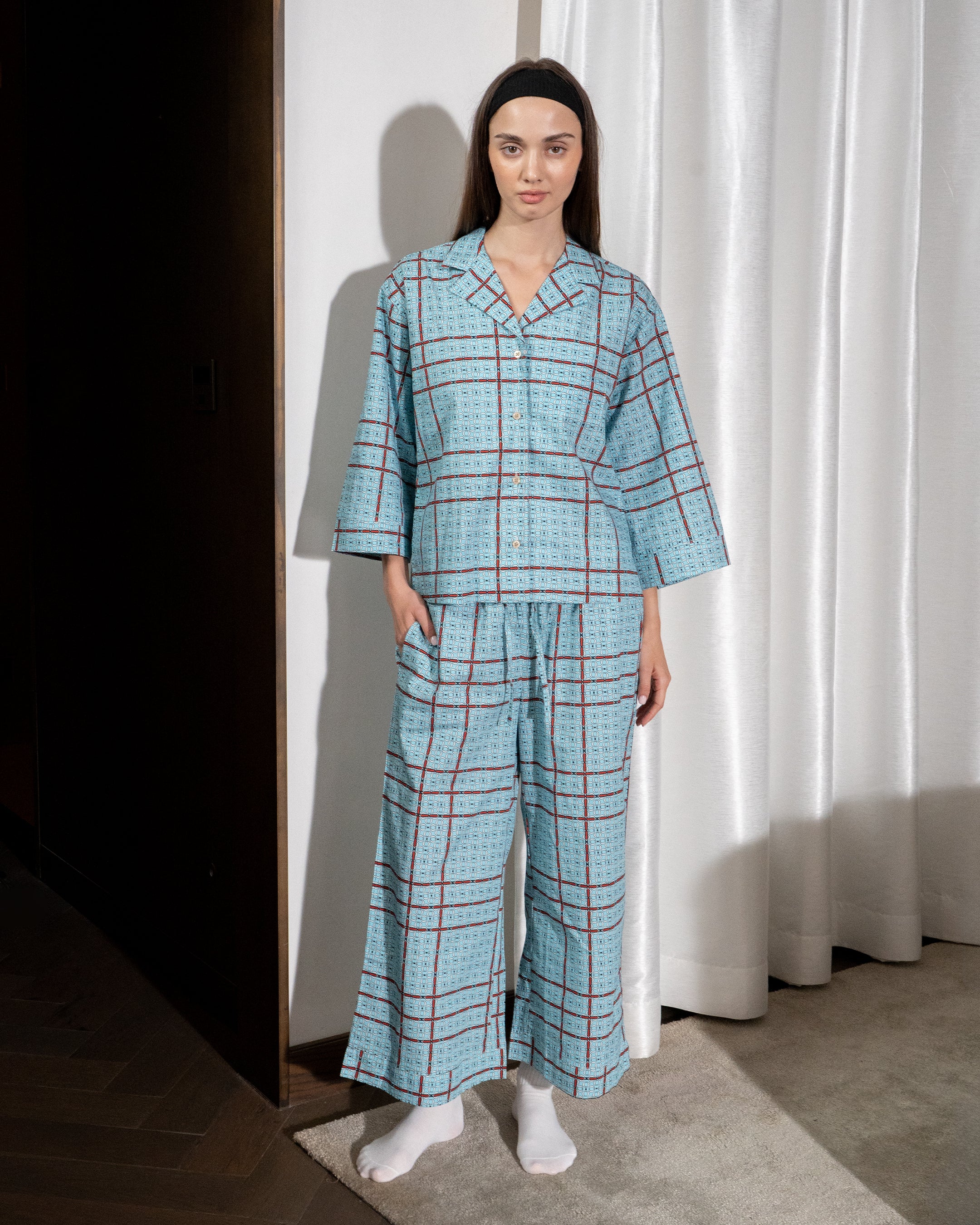 Woman wearing a blue checkered pajama set standing against a white curtain.